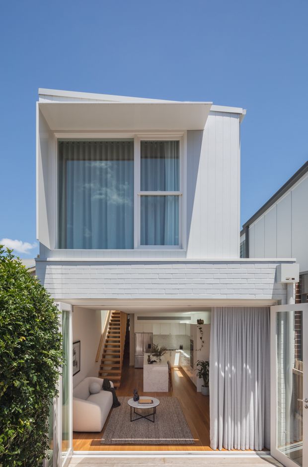 A bright, narrow multi-storey townhouse facade with white vertical cladding, white-painted brickwork and a projecting upper-level window canopy. The ground floor is open to a contemporary living room and kitchen, visible through large glazed doors, with timber flooring, a white sofa, island bench, staircase and full-height curtains. The exterior has a clean, compact urban character under a clear blue sky.