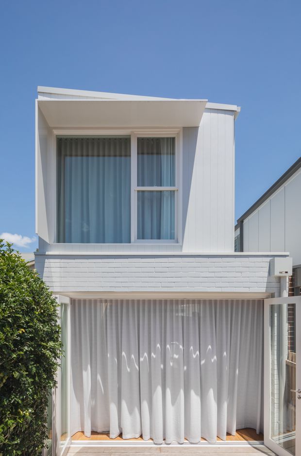 Front exterior view of a narrow, multi-storey residential townhouse. The facade is predominantly white, with a projecting upper-level box containing large glazed windows, vertical white cladding, and a low-pitched roofline. The lower level has a broad glazed opening screened by sheer curtains, while a small leafy tree and neighbouring building edge frame the facade.