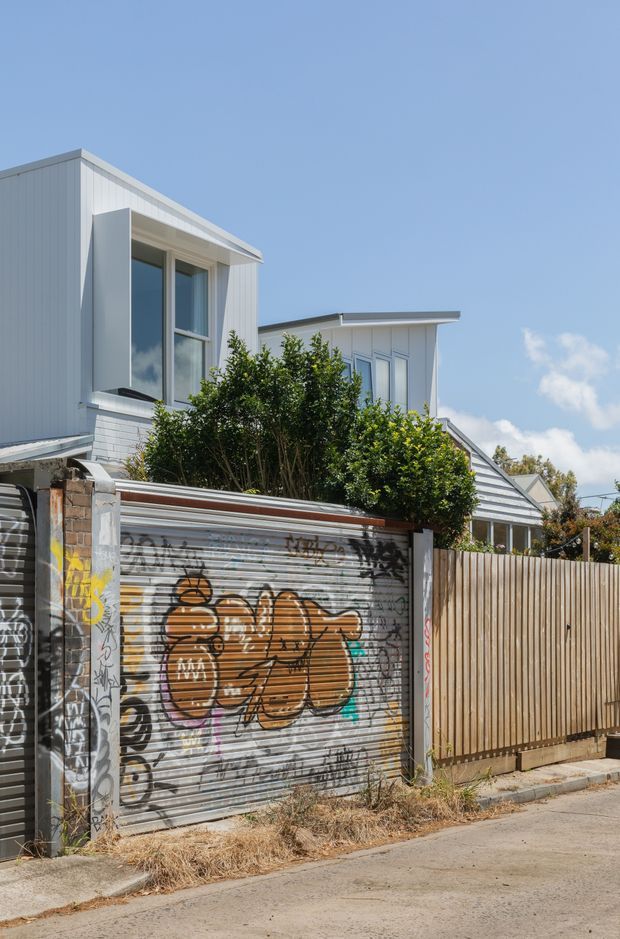 Street-facing view of a contemporary multi-storey residential building with white vertical cladding, projecting windows, and a low-pitched roof. The property is enclosed by a tall timber privacy fence and a graffiti-covered roller garage door, with mature shrubs screening the facade and a concrete-paved street in the foreground.