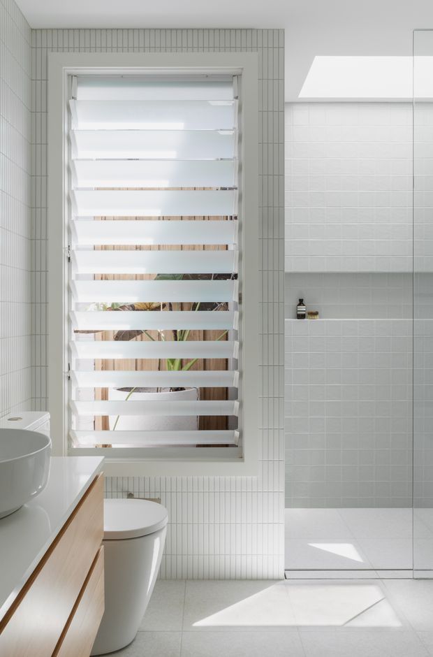 Bright contemporary bathroom with white small-format tiled walls, a large louvred window with filtered daylight, a frameless glass shower enclosure, white toilet and basin, and a light timber vanity. The room has a clean, airy, minimalist appearance with strong natural light and subtle greenery visible through the window.
