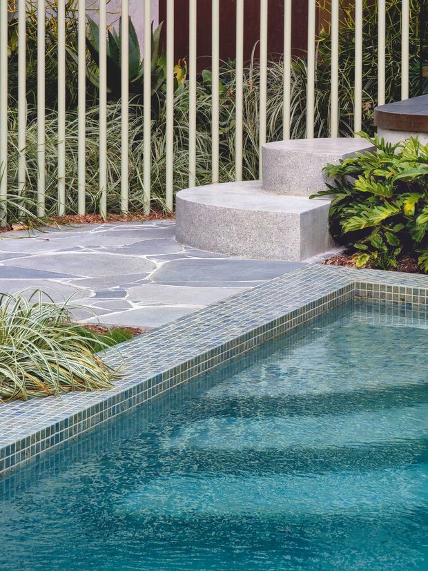 Close-up view of a landscaped outdoor pool area with turquoise water, a small green mosaic-tiled pool edge, irregular grey stone paving, curved concrete steps, tropical planting, and a vertical metal garden screen.