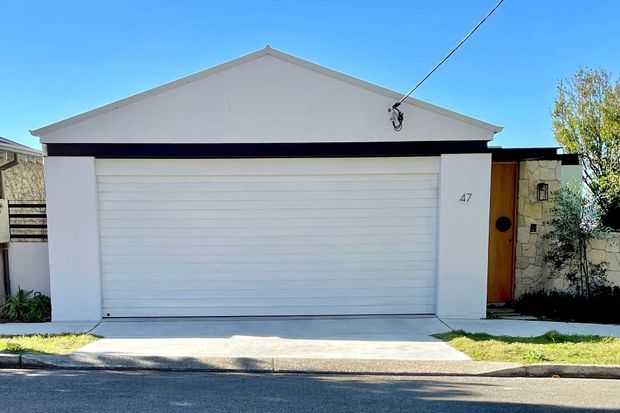 Street-facing view of a compact single-storey residential garage and entry facade. The building has a white rendered gable-front form, a wide white horizontal-panel garage door, a black fascia band, and a timber pedestrian entry door beside a textured stone wall. A concrete driveway and small landscaped margins sit directly in front.