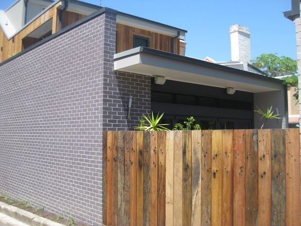 Street-facing view of a compact contemporary multi-level residence. The facade combines dark grey brickwork, warm vertical timber cladding, a flat-roofed projecting carport or garage canopy, dark-framed glazing, and a tall timber privacy fence. A white masonry chimney and neighbouring buildings are visible in the background.