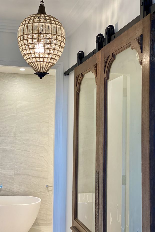 A refined bathroom interior featuring a freestanding white bathtub, pale stone-look wall finish, and a large antique-style timber-framed mirrored barn door on black sliding hardware. A prominent beaded glass pendant light adds a decorative focal point, while the overall palette is light and warm with dark timber and metal accents.