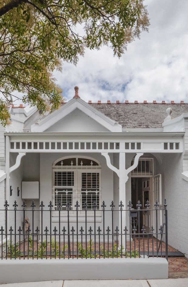 Front exterior view of a restored-looking white heritage terrace or cottage, featuring a steep gabled roof, decorative timber verandah with fretwork and curved brackets, ornate finials, sash windows, a traditional doorway, brick-paved entry and black wrought-iron fencing. Mature tree branches frame the upper portion of the image.