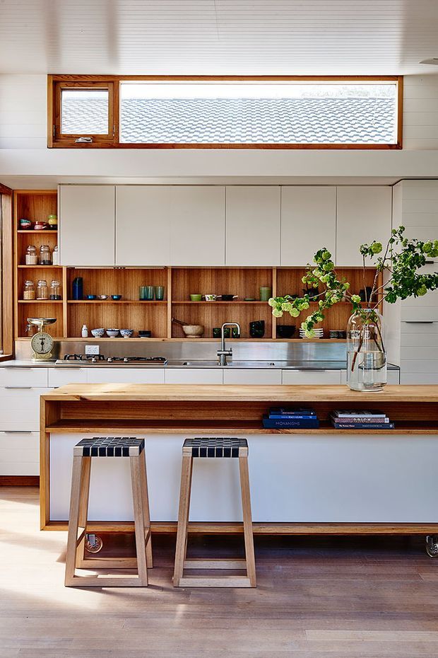 Bright, contemporary kitchen with a long island featuring a timber benchtop, white panelled cabinetry, open timber shelving, and integrated stainless-steel appliances. Two timber bar stools sit at the island, while a high horizontal timber-framed window brings in natural light. Dark timber flooring and a large glass vase with leafy branches add warmth and contrast.