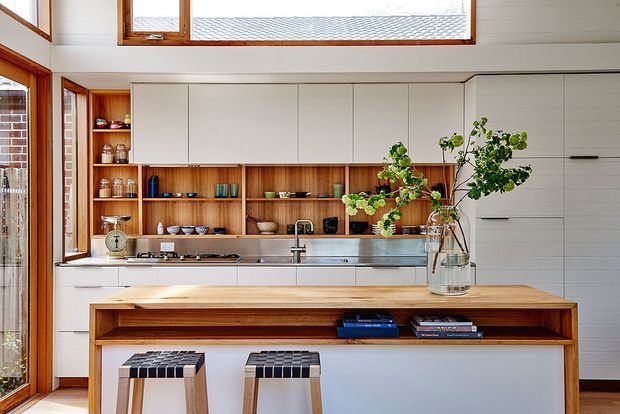 Bright contemporary kitchen with white flat-panel cabinetry, warm timber open shelving and island cabinetry, stainless-steel appliances and benchtop, and timber-framed windows. Two stools sit at the large island, with natural greenery adding a soft accent.