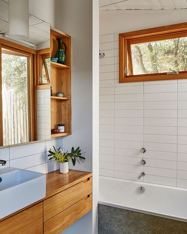 Bright bathroom interior featuring a white tiled shower and bath area, timber-framed windows, a timber vanity with integrated drawers, a white basin, mirrored wall, and open timber shelving with decorative objects. Natural greenery is visible outside the windows, while a pendant light and potted plant add warmth.
