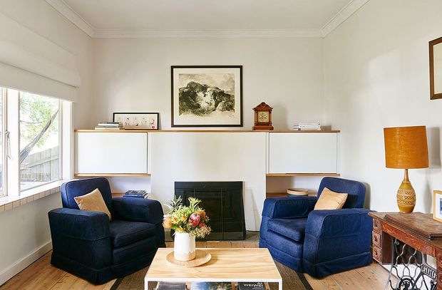 A bright, traditional living room with two deep navy upholstered armchairs facing a low timber coffee table and a dark tiled fireplace. Built-in white wall cabinetry with open timber shelves lines the wall, complemented by framed artwork, a vintage mantel clock, a warm table lamp, and a timber console. Large windows bring in abundant natural light, while decorative cornices and neutral walls add classic character.