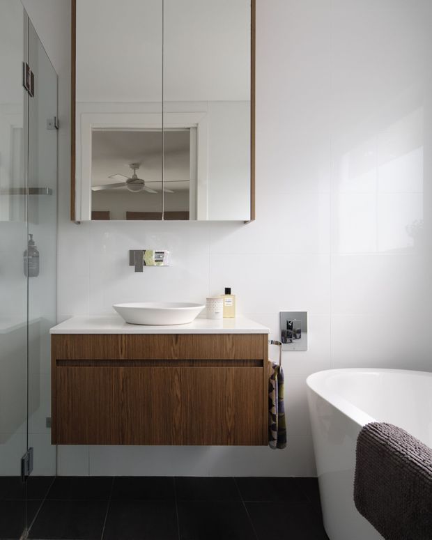 Contemporary bathroom featuring a floating dark timber vanity with a white vessel basin, mirrored wall cabinet, white tiled walls, black floor tiles, frameless glass shower, and a freestanding white bathtub. The room is bright and clean with restrained modern detailing.