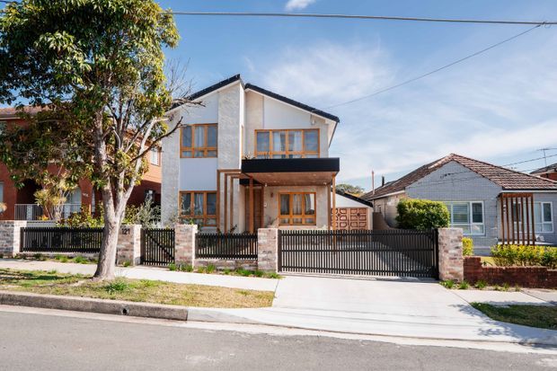 Street-facing view of a contemporary two-storey residential house with a white rendered facade, dark pitched roof, warm timber-framed windows, projecting dark balcony, front fence and driveway. The home sits in an established suburban streetscape alongside neighbouring houses and mature trees.