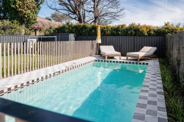 A compact rectangular swimming pool in a fenced residential backyard, edged with patterned grey-and-white paving and a contrasting tiled waterline. Two outdoor loungers and a closed umbrella sit at the far end, with hedges, mature trees and neighbouring residential elements beyond the dark privacy fencing. The scene is bright and sunny with a clean, resort-like outdoor atmosphere.