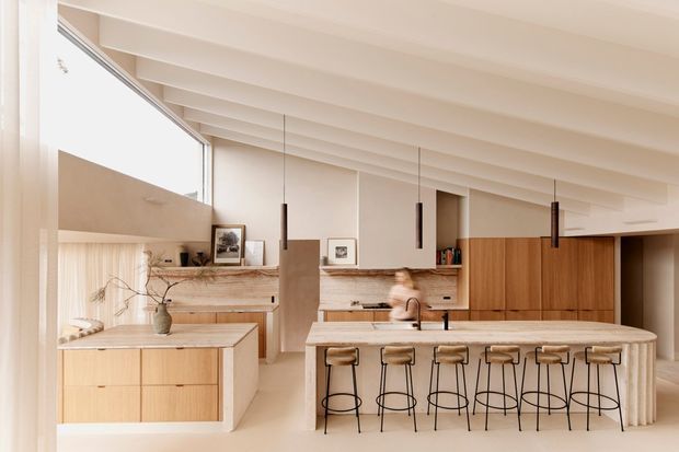 Bright contemporary kitchen with a large central island and six stools, pale timber cabinetry, a matching timber and stone-look benchtop, integrated appliances, open shelving, and a warm textured splashback. Exposed painted ceiling beams, floor-to-ceiling glazing, and slim black pendant lights add architectural character.