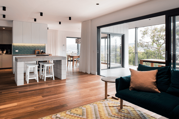 A bright open-plan interior combining a contemporary kitchen, dining area and living room. The kitchen features white built-in cabinetry, a tiled island, white bar stools and a green tiled splashback. Warm timber flooring extends through the space, with a dark teal sofa, patterned rug and timber coffee table in the living area. Large black-framed sliding glass doors open to a balcony or terrace, bringing in abundant natural light.