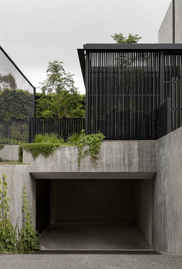 Exterior architectural view of a contemporary concrete residence, showing a recessed open garage or undercroft at ground level and an upper terrace enclosed by black vertical timber or metal screening. Raw grey concrete walls, dark framing, planted edges, and a chain-link fence create a restrained, utilitarian composition integrated with dense greenery.