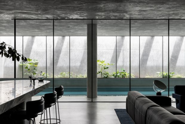 A dramatic, contemporary open-plan interior with a long polished stone island or bar on the left, black stools, a dark upholstered sofa in the foreground, and a low teal built-in seating or joinery element along the window wall. Full-height black-framed glazing spans the rear of the room, with indoor planting and strong diffused daylight. The ceiling and structural column have a raw, dark concrete-like finish, creating a refined industrial atmosphere.