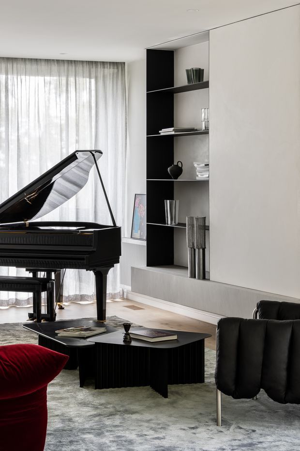 Contemporary living room with a black grand piano, sculptural black coffee table, black leather seating, a red upholstered chair, sheer full-height curtains, and a recessed open display shelving unit integrated into a pale wall. The room has a refined monochromatic palette with subtle warm timber flooring and a large grey rug.