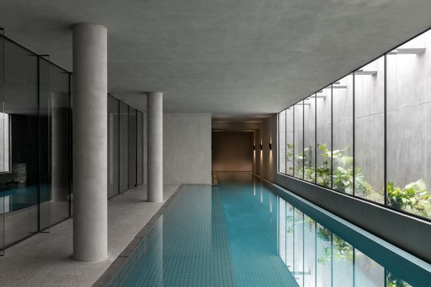 A long, narrow indoor swimming pool occupies a stark modern concrete space. The pool is bordered by large floor-to-ceiling glazed walls on one side, revealing a planted lightwell or courtyard, while cylindrical concrete columns and dark glass partitions line the opposite side. The restrained palette and low ceiling create a calm, gallery-like atmosphere with strong linear perspective.