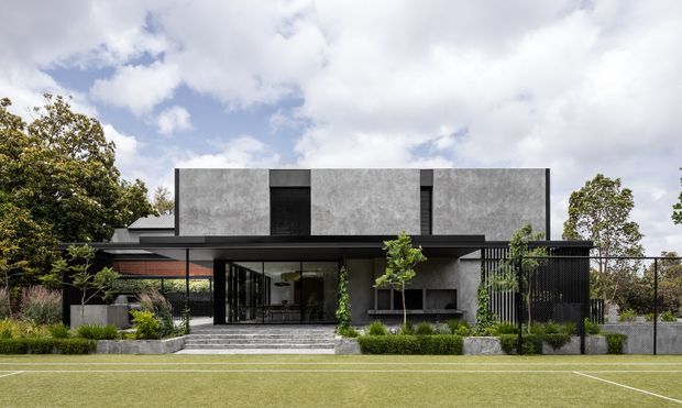A contemporary two-storey residential building viewed from the front across a marked sports field. The home has a rectilinear concrete or rendered upper volume, extensive dark-framed glazing, a broad flat-roofed ground-floor canopy, dark vertical screening, landscaped planting, and a stepped entry. The composition is strongly modern and geometric, with muted grey, black, and green tones.