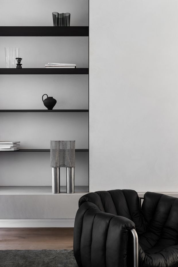 A moody, minimalist living room vignette featuring a black leather lounge chair in the foreground, a built-in open shelving unit with dark shelves and neutral plaster backing, curated monochrome ceramics and books, and a sculptural metallic table lamp. The room has dark timber flooring, a grey rug, and a restrained grey, black, and metallic palette.