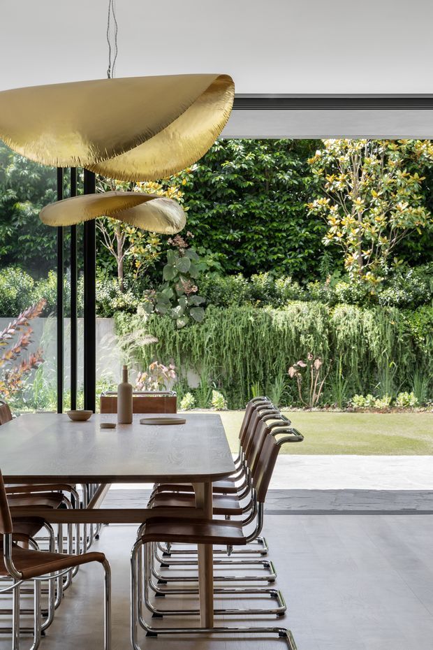 A refined dining area opening onto a landscaped garden through large black-framed glazed doors. A long light stone or concrete dining table is surrounded by timber and chrome cantilever chairs, with sculptural gold pendant lights overhead. Pale large-format flooring, dark window framing, lush planting, and a seamless indoor-outdoor connection create a contemporary, elegant setting.