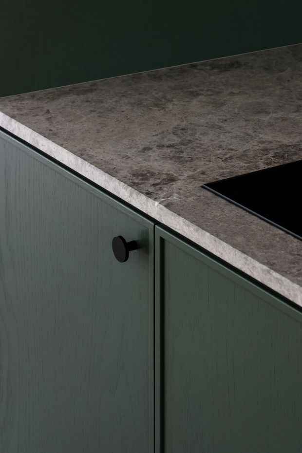 Close-up detail of dark green vertically textured kitchen cabinetry with a small black round knob, paired with a dark grey-brown veined stone benchtop and a partially visible black cooktop. The image focuses on refined fixed joinery and benchtop materials, with a dark, moody contemporary aesthetic.