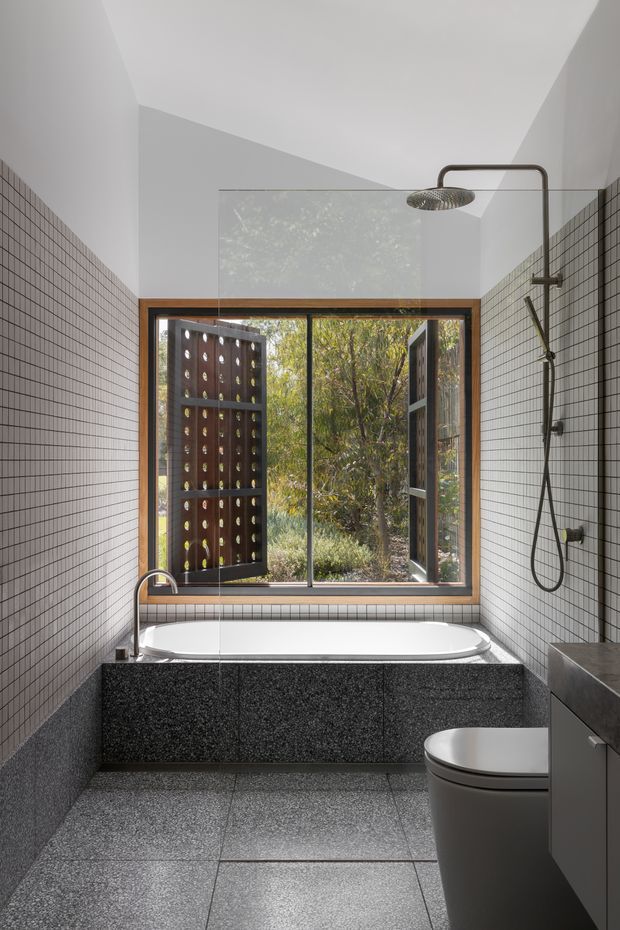 A contemporary bathroom with a freestanding bathtub beneath a large timber-framed window, black and white square wall tiles, dark terrazzo-style wall and floor surfaces, and a wall-mounted shower with ceiling rain shower. The window opens toward a landscaped garden and screened outdoor area, bringing in abundant natural light. A toilet and dark vanity are visible in the foreground.