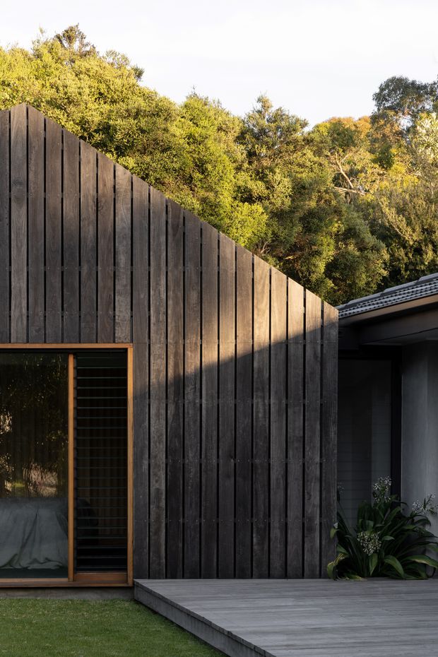 Exterior view of a contemporary house surrounded by mature vegetation. The facade is clad in dark vertical timber boards forming a tall angled screen, with a large glazed opening and timber-framed louvred door. A grey timber deck leads to a recessed entry or covered outdoor area, with a tiled roof edge visible at right.