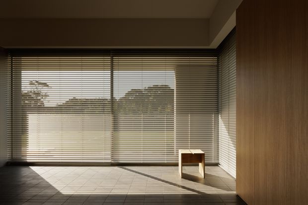 A minimalist, mostly empty interior with full-height corner glazing fitted with horizontal blinds. Warm sunlight casts strong linear shadows across a large tiled floor. A small simple wooden stool sits near the windows, while a full-height warm timber-clad wall or built-in panel occupies the right side. The space has a calm, restrained and architectural character.
