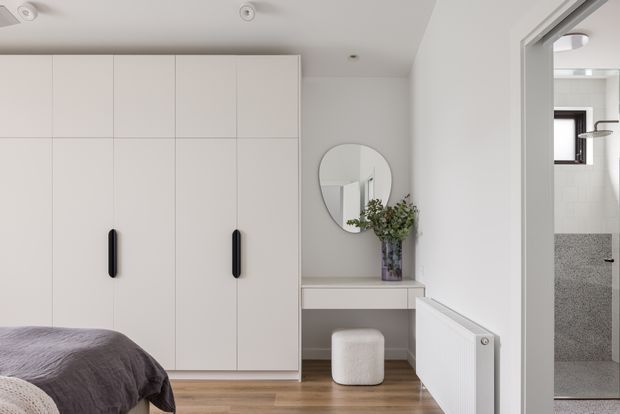 A bright, minimalist bedroom with full-height white built-in wardrobes, a built-in dressing console and round mirror, timber flooring, and a partially visible upholstered bed. A bathroom is visible through the open doorway, with a wall-mounted shower and grey textured wall finish.