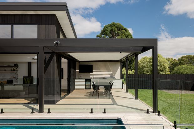 A contemporary residential outdoor entertaining area with a covered pergola beside a swimming pool. The space includes an outdoor kitchen with white cabinetry, a dining table, dark metal framing and screening, timber-look decking, pale paving, and clear glass pool fencing. The modern house and landscaped lawn are visible around the terrace.