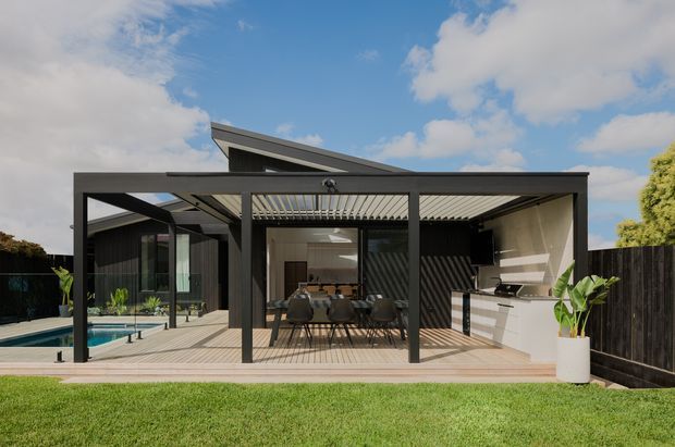 Contemporary single-storey home viewed from the backyard, featuring a dark-framed covered outdoor entertaining area with a slatted pergola roof, dining setting, and compact outdoor kitchen. A timber deck surrounds the entertaining zone and pool, with dark fencing and landscaped lawn.