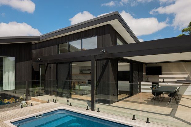 Contemporary multi-level home exterior viewed from a poolside timber deck, featuring dark vertical cladding, expansive glazing, a projecting roofline, and a covered outdoor dining and kitchen area. The image also shows a swimming pool, glass balustrades, and a bright blue-sky setting.