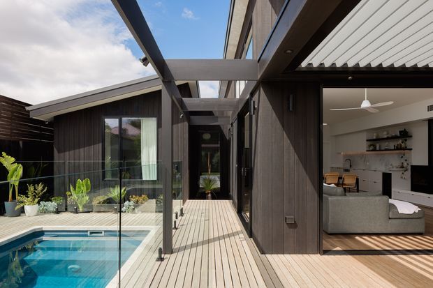 Contemporary courtyard-style residential exterior with a timber deck walkway beside a small swimming pool, dark vertical cladding, extensive glazing, and a covered outdoor connection between wings. An open living room with a sofa, shelving, dining area, and kitchen is visible through the glazing on the right. The scene has a strong indoor-outdoor connection, exposed steel or painted structural beams, and a light timber-lined soffit.
