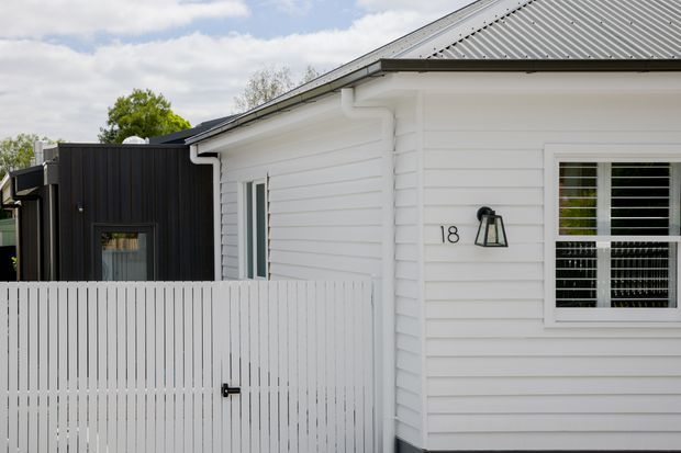 Exterior view of a single-storey residential building with white horizontal weatherboard cladding, a grey corrugated pitched roof, white framed windows with horizontal security or privacy bars, and a white vertical-slat gate. A contrasting black-clad extension or adjoining section is visible on the left, with the house number and wall-mounted light on the facade.