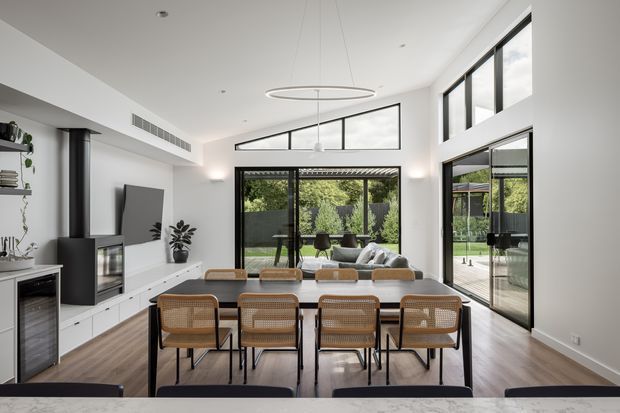 Bright open-plan dining and living area with a long timber dining table and woven cane dining chairs in the foreground. White built-in cabinetry, a television, shelving and a compact bar refrigerator line the left wall, while expansive black-framed glazing and sliding doors open to a green outdoor area. A high white ceiling with a suspended circular pendant gives the room an airy contemporary feel.