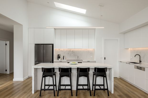 Bright contemporary white kitchen with a large marble-look island and seating for four, streamlined white cabinetry, integrated appliances, under-cabinet lighting, black pendant and hardware accents, timber-look flooring, and a skylight.