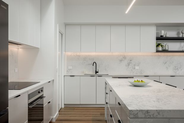 Contemporary white kitchen with full-height handleless cabinetry, a large veined stone island, matching splashback, integrated appliances, black tapware, open display shelving, and warm timber flooring. A linear ceiling light provides understated illumination.