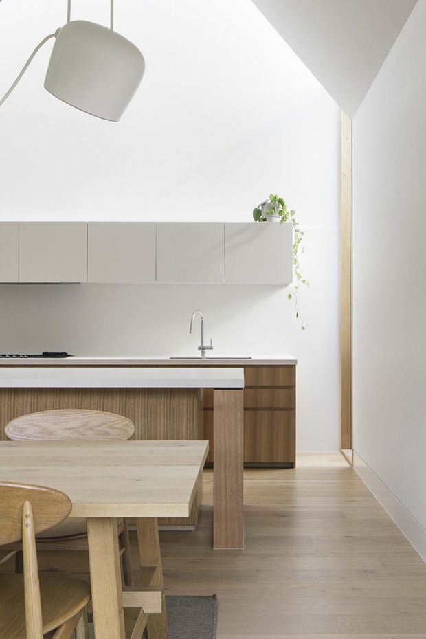 Bright, minimalist kitchen-dining interior with pale wood flooring and cabinetry, a long light timber dining table with rounded wooden chairs, and a compact kitchen featuring smooth matte light-grey overhead cabinets, timber base cabinetry, a slim benchtop, integrated cooktop, and stainless-steel tap. White walls and ceiling create an airy setting, with a large sculptural pendant light and a trailing potted plant adding subtle detail.