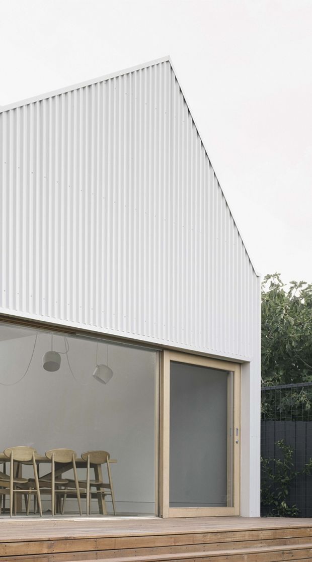 Exterior view of a compact contemporary building with a steep gable-like roof profile and white vertically ribbed metal cladding. Large glazed openings reveal a simple dining area with timber chairs and pendant lights, while a timber deck and steps occupy the foreground. The restrained white, grey, and natural timber palette creates a clean modern appearance.
