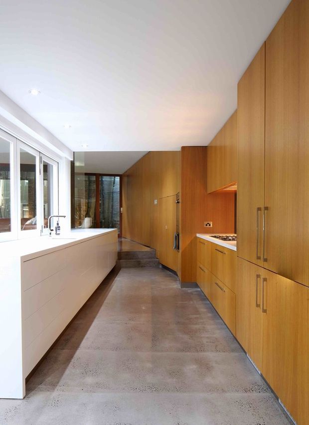 A narrow, contemporary kitchen with extensive warm timber cabinetry along the right wall, integrated appliances, a white island or peninsula with a sink beside large glazed doors, and a polished concrete floor. Steps lead toward a darker adjoining area at the rear. The room is bright and minimalist with clean linear detailing.