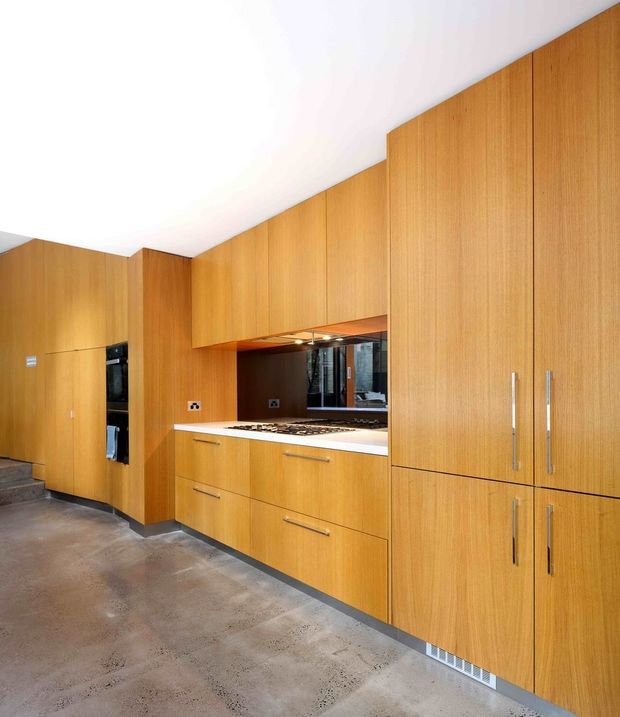 A contemporary kitchen with extensive warm timber cabinetry, integrated tall storage, a built-in oven, gas cooktop, pale benchtop and dark splashback. The cabinetry runs along multiple walls, and the room has a polished concrete floor with a clean, minimalist fit-out.
