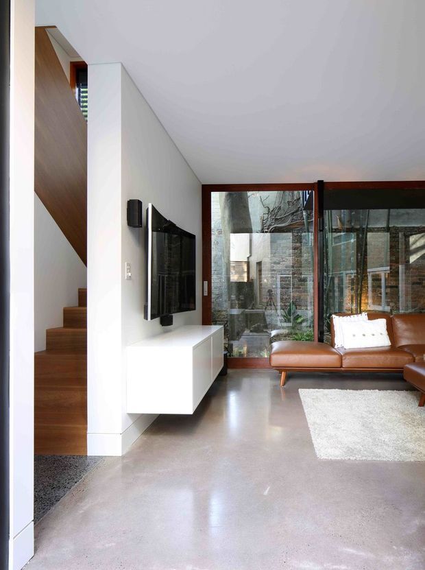 A bright contemporary living room with polished concrete flooring, white walls, and a sculptural timber staircase rising along the left side. A wall-mounted television sits above a floating white media cabinet, while a brown leather sofa faces the screen. Full-height dark timber-framed glazing opens views to a planted courtyard and masonry wall outside. The room has a clean, minimalist architectural character with strong indoor-outdoor connection.