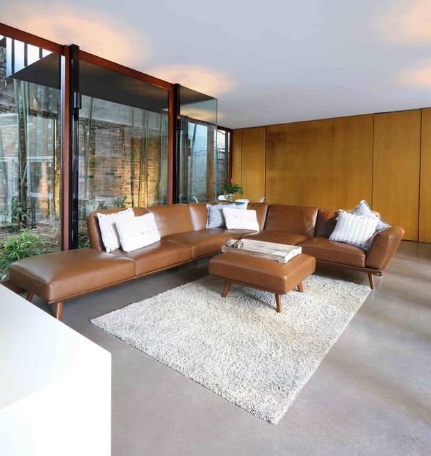 A spacious, refined living room with a large cognac-brown leather sectional and matching ottoman arranged over a textured cream rug. Full-height glazing opens views to a brick and planted courtyard, while warm timber wall panelling and dark-framed glass doors create a mid-century-inspired interior with strong indoor–outdoor connection.