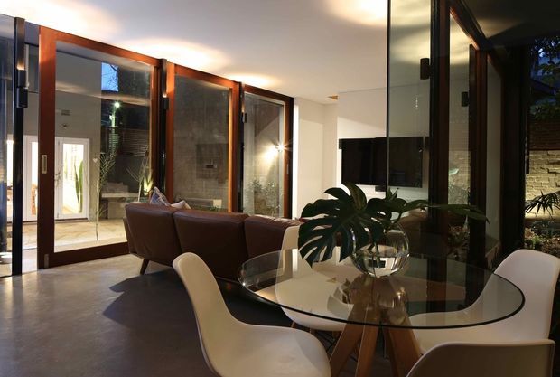A contemporary open-plan living and dining area with extensive dark timber-framed glazing and folding doors opening to a planted courtyard. The room features a glass dining table with sculptural white chairs, a brown leather sofa, polished dark flooring, and a wall-mounted television. Indoor plants and reflected courtyard views add a warm connection to the outdoors.