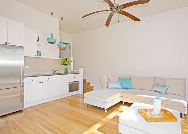 Bright, open-plan living area with a compact white kitchen along the left wall and a large light upholstered sectional sofa in the foreground. The room has honey-toned timber flooring, white walls and cabinetry, stainless-steel appliances, turquoise pendant lights and cushions, and a prominent wood-bladed ceiling fan. A short set of timber steps leads upward from the living area.