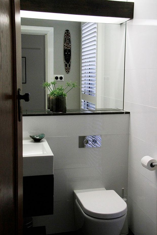 Compact contemporary bathroom featuring a wall-hung toilet, floating dark vanity with integrated white basin, large mirror, pale wall tiles, black stone-look ledge, and white louvre window. Dark timber door framing and minimal decorative accents add contrast.