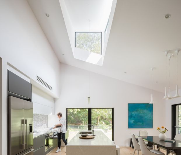 Bright, contemporary open-plan kitchen and dining area with a high ceiling and large skylight. The kitchen features a long white island with an integrated sink, dark lower cabinetry, a stainless-steel refrigerator, and a reflective mosaic-tile splashback. Floor-to-ceiling glazing and black-framed windows overlook greenery. A dining table with upholstered chairs, sculptural pendant lights, and a blue abstract artwork occupy the adjoining dining zone.