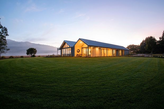 A low, elongated contemporary building set in a broad manicured lawn at dusk. The structure has a dark pitched metal roof, warm timber-toned illuminated interiors, extensive floor-to-ceiling glazing, and a glazed gabled end. Trees, distant hills, and a calm body of water appear in the background.