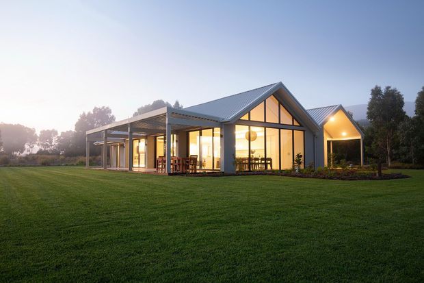 A contemporary single-storey house set within a broad lawned landscape at dusk. The building has multiple steep gabled volumes with dark metal roofing, extensive floor-to-ceiling glazing, light-coloured structural framing, and a covered outdoor terrace or verandah. Warm interior lighting glows through the glass, with dining and living areas visible inside. Mature trees and a softly misty rural-looking setting surround the home.